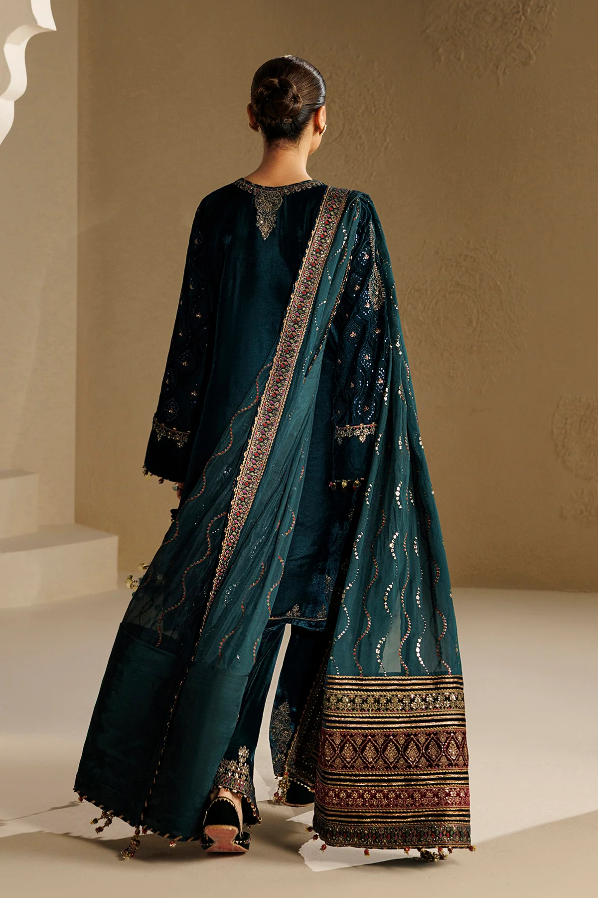 A woman shown from the back wearing a dark teal traditional outfit with metallic embroidered patterns and a matching draped scarf, photographed in an indoor studio setting with warm beige walls.