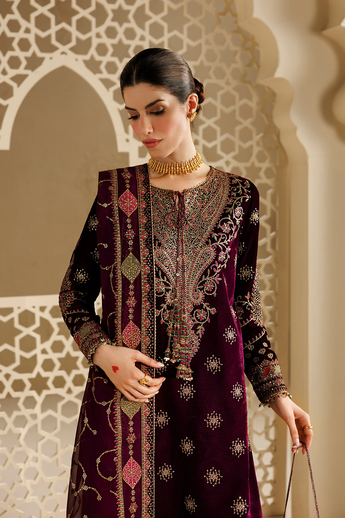 A woman models a deep purple velvet traditional South Asian outfit with intricate gold, pink, and green embroidery, standing in front of a beige archway with a geometric screen.