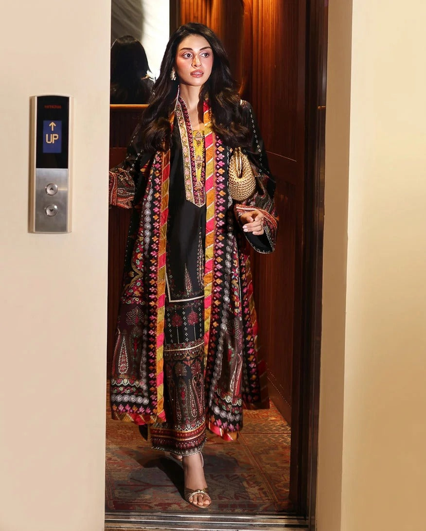 A woman wearing a traditional South Asian outfit with a long multicolor printed outer robe over a black kurta and matching trousers, standing in an elevator.