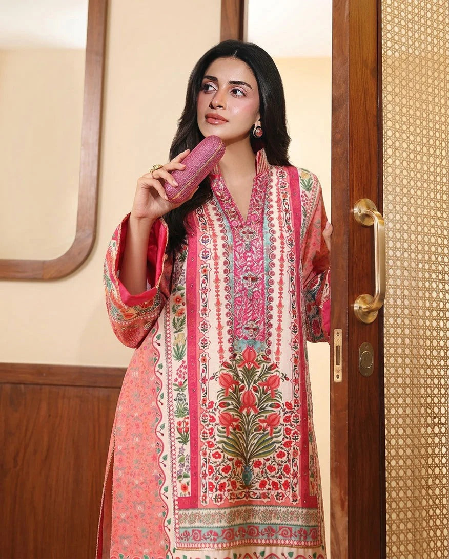 A woman wearing a pink and red printed traditional kurta with floral motifs, holding a matching pink clutch in an indoor setting with wooden door frames.