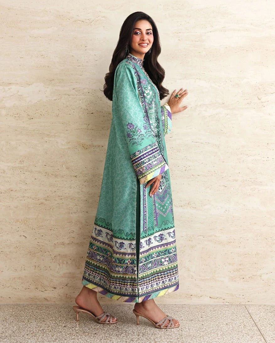 A woman modeling a long mint green traditional dress with intricate multicolor embroidery and a patterned border, standing against a textured beige stone wall.