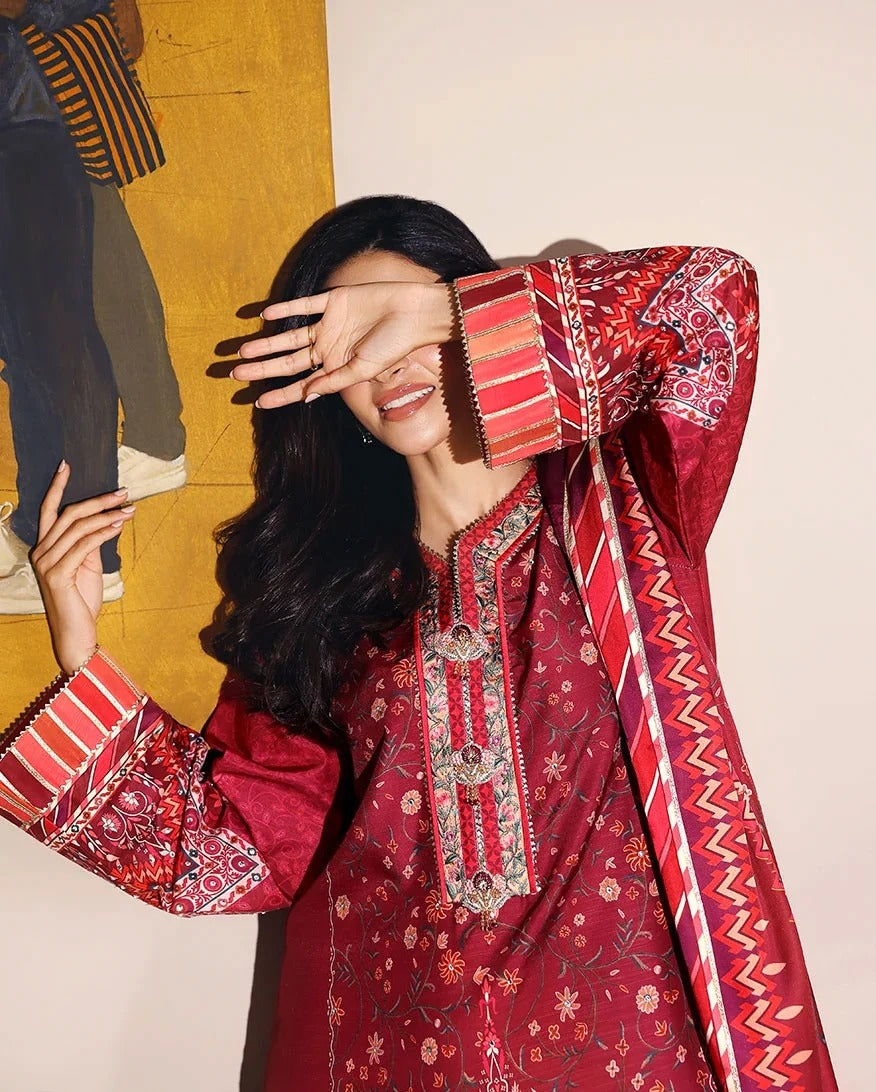 A woman wearing a red and gold embroidered traditional kurta with a matching patterned dupatta, posing playfully with her hand over her eyes against a beige wall and a yellow art print.