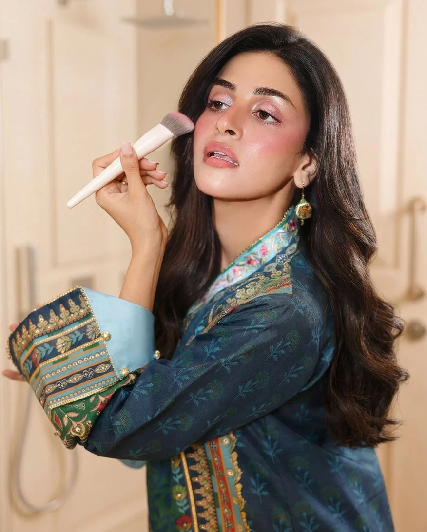 A woman with long dark hair applies makeup with a brush while wearing a teal embroidered traditional outfit with a light blue inner layer and ornate gold and green patterns on the sleeve cuffs.
