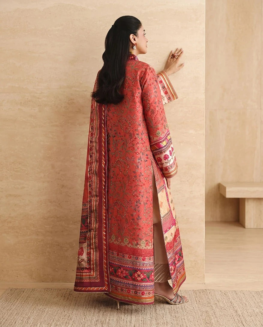 A woman is shown from the back wearing a long red printed traditional outfit with ornate multicolor borders and sleeve patterns, standing in a minimalist beige interior setting.