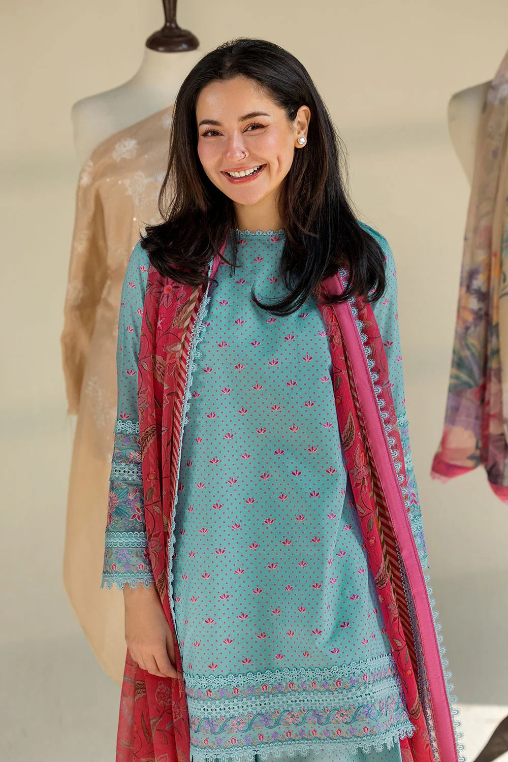 Woman wearing a teal embroidered outfit with a pink dupatta, standing in front of mannequins.