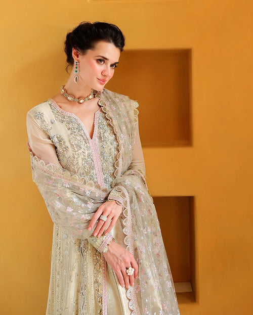 A woman models a traditional South Asian outfit featuring a light beige/cream embroidered kurta with a matching sheer dupatta, set against a warm ochre-colored wall with recessed panels.