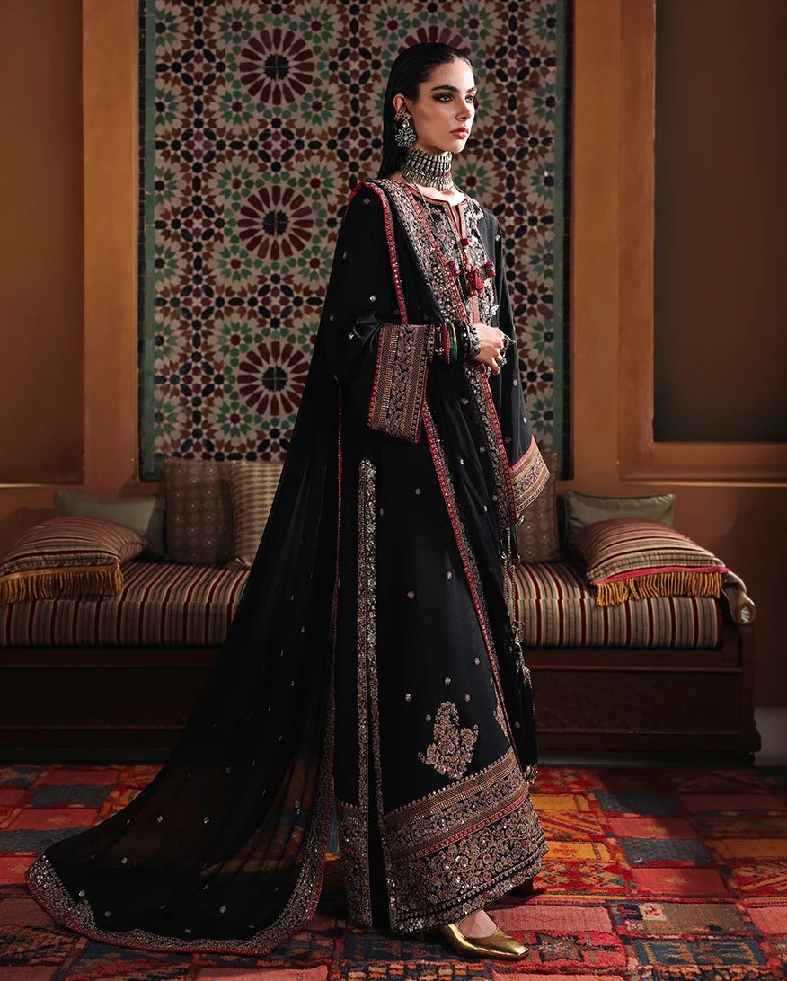 A woman models a traditional black embroidered ethnic outfit with a long dupatta in an ornate indoor setting featuring a patterned wall hanging and cushions.