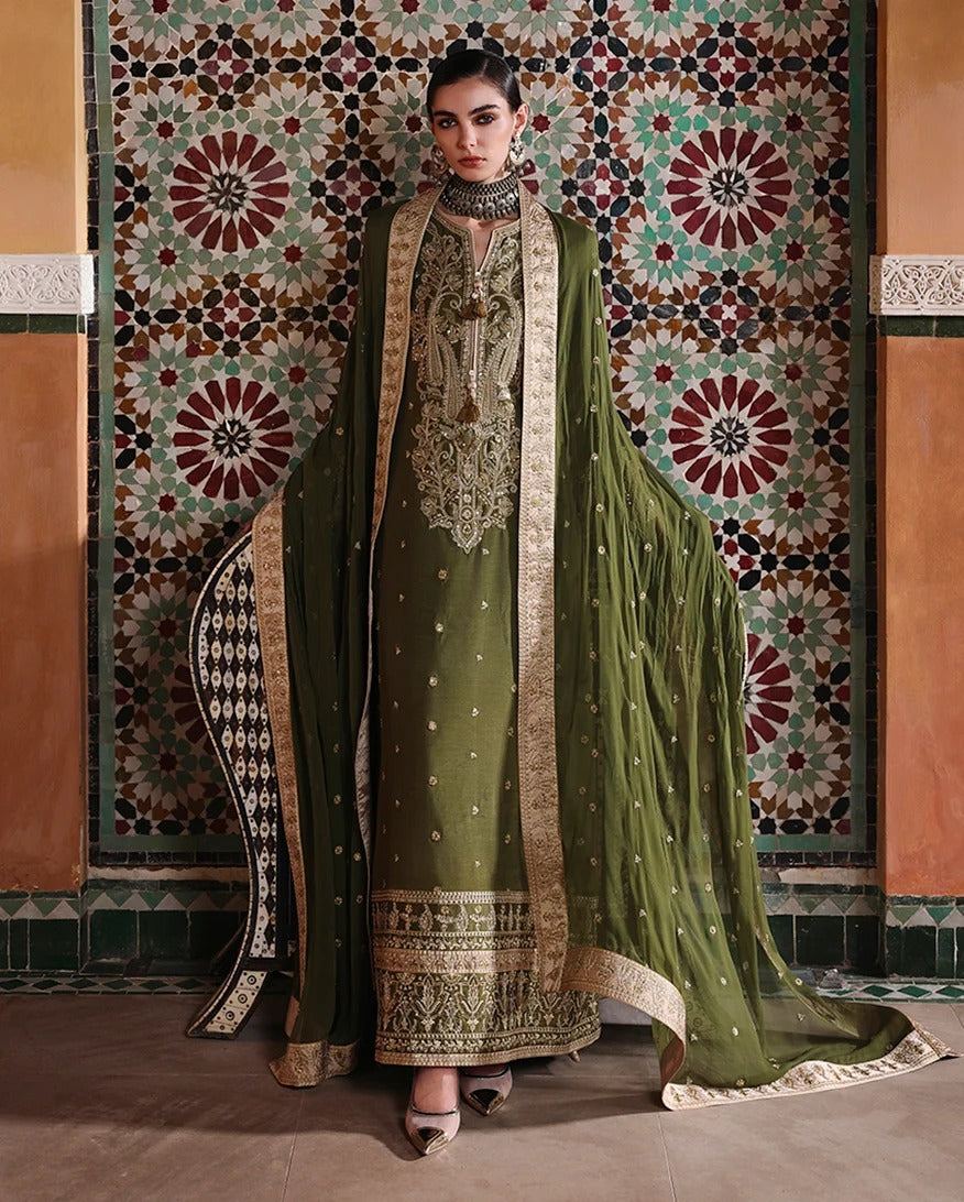 A woman models an olive green traditional South Asian salwar kameez suit with intricate gold embroidery, a matching dupatta, and a high-neck choker, posed in front of an ornate, patterned tile wall.