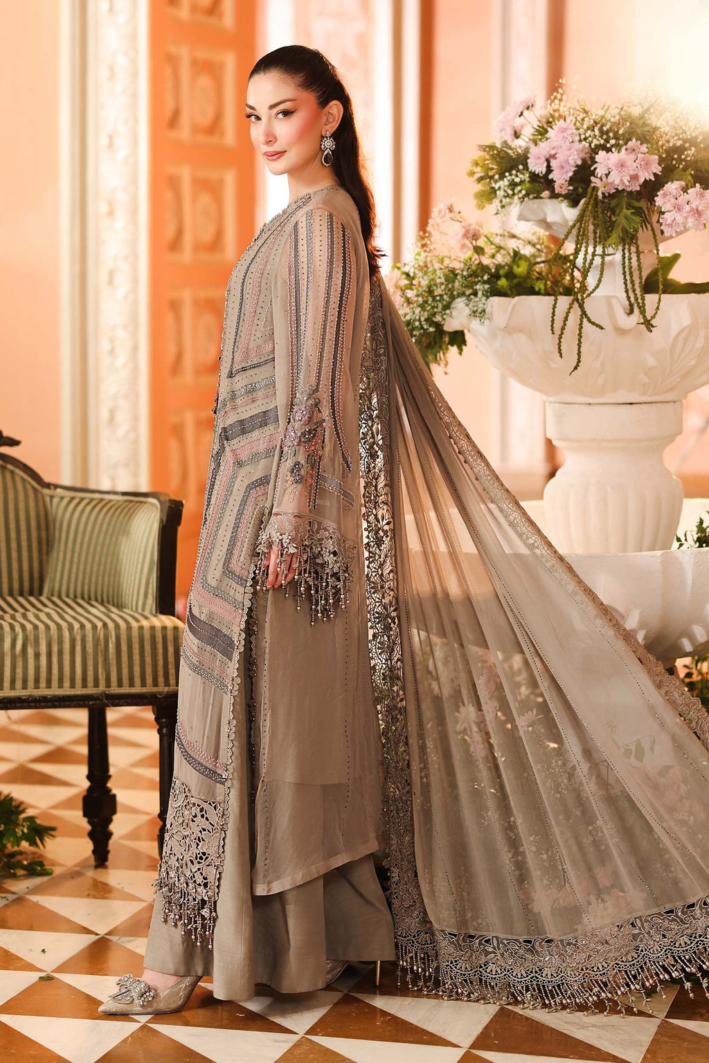A woman models a traditional South Asian salwar kameez outfit in a warm, elegant room. The outfit consists of a taupe-beige long-sleeved kameez jacket with intricate silver and gold geometric embroidery, matching wide-leg trousers, and a sheer dupatta with floral embroidery and a silver border.