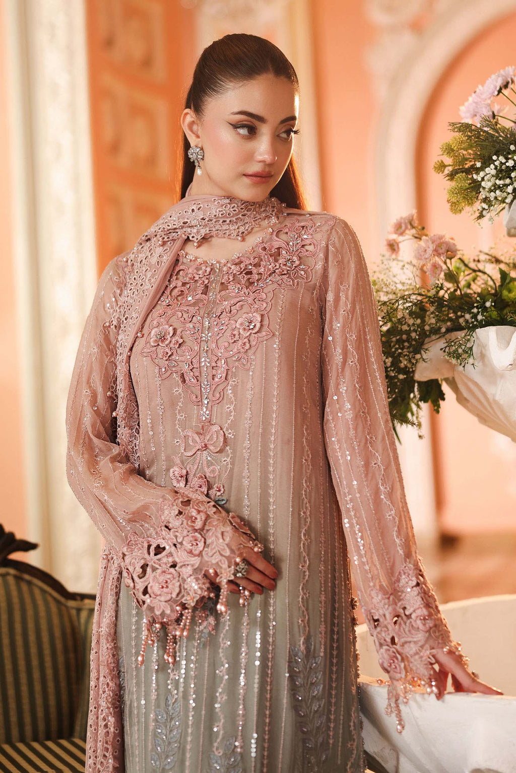 A woman wearing a heavily embellished light pink traditional outfit with sequins, embroidery, and floral lace details, posed indoors against a warm peach-toned background with flowers and furniture partially visible.