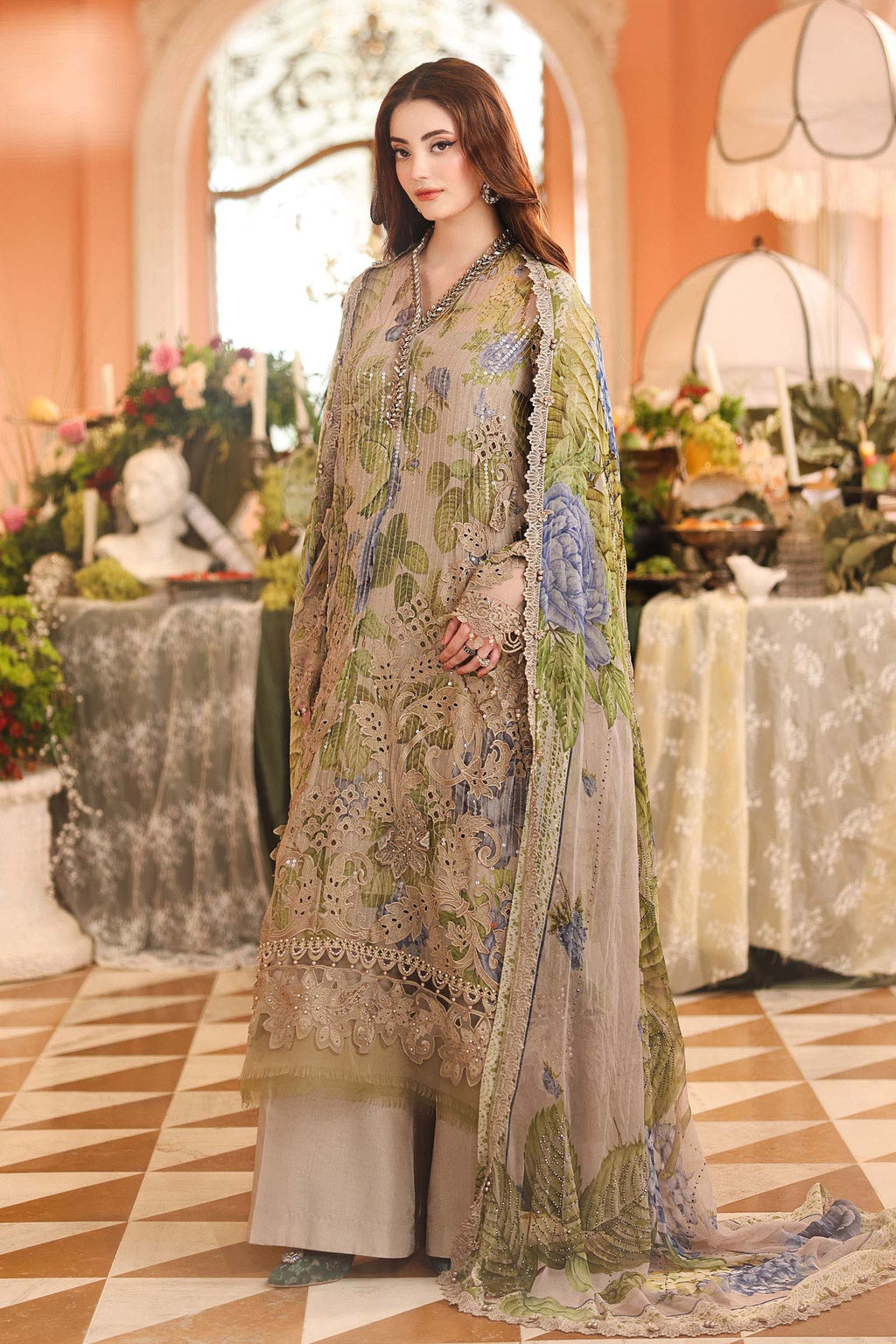 A woman models a traditional three-piece salwar kameez suit in a warm, decorated indoor setting. The outfit is a light beige/taupe color with intricate green and blue floral embroidery, a matching embroidered dupatta, and light beige trousers.