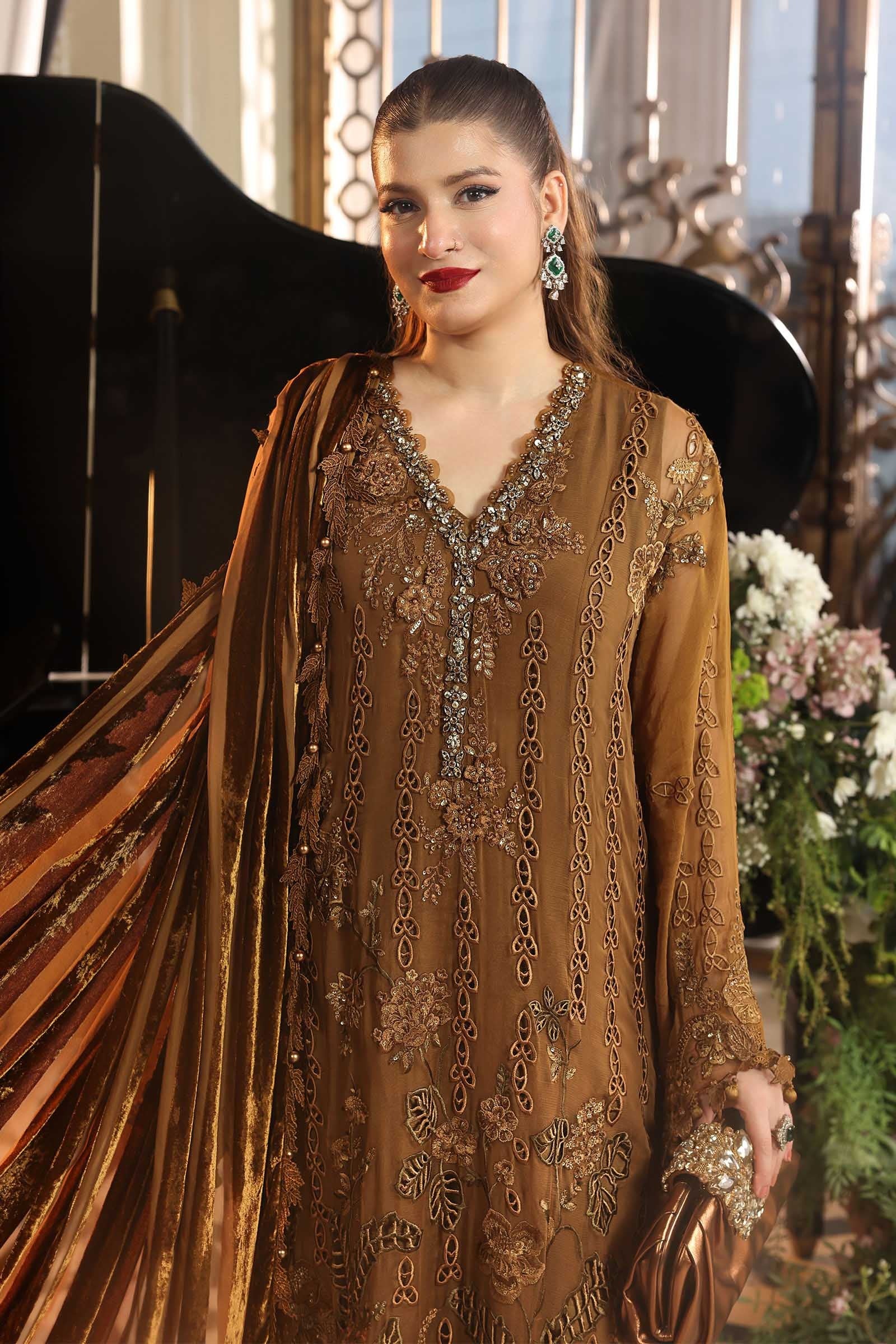 A woman models a brown embroidered traditional outfit with a matching draped scarf, featuring intricate metallic embellishments and a V-neckline, set against an elegant indoor background with a piano and floral arrangements.