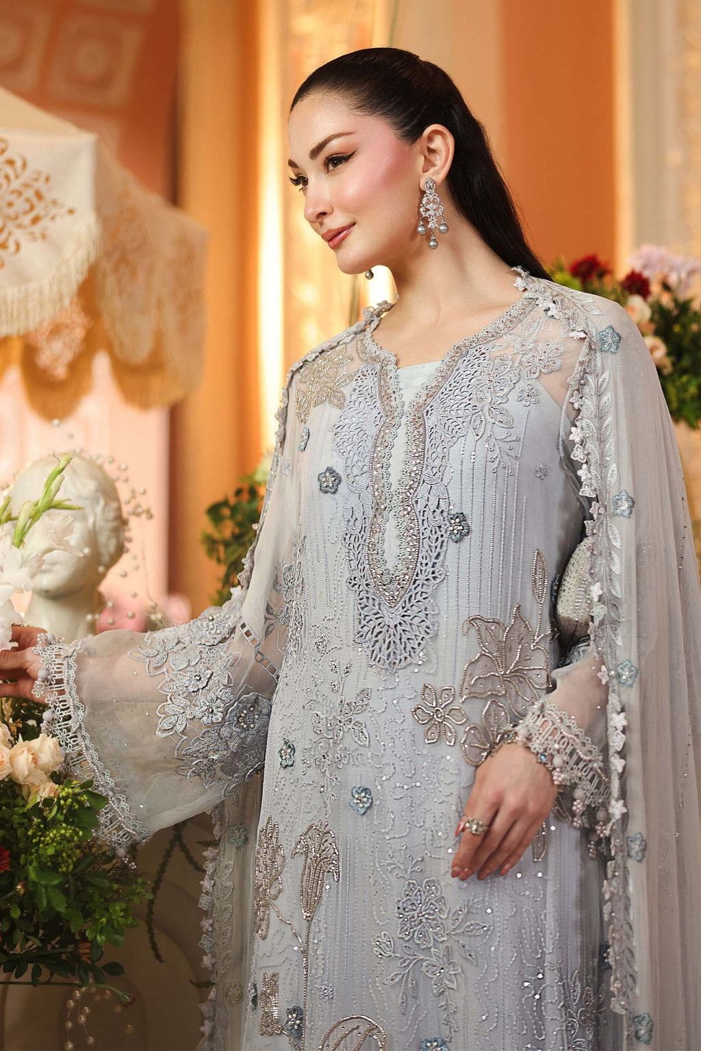 A woman wearing a light silver-gray embroidered traditional outfit with sheer sleeves and floral detailing, posed in an elegant indoor setting with warm beige walls and decorative flowers.