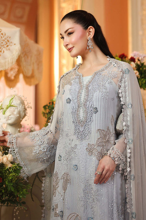 A woman wearing a light silver-gray embroidered traditional outfit with sheer sleeves and floral detailing, posed in an elegant indoor setting with warm beige walls and decorative flowers.