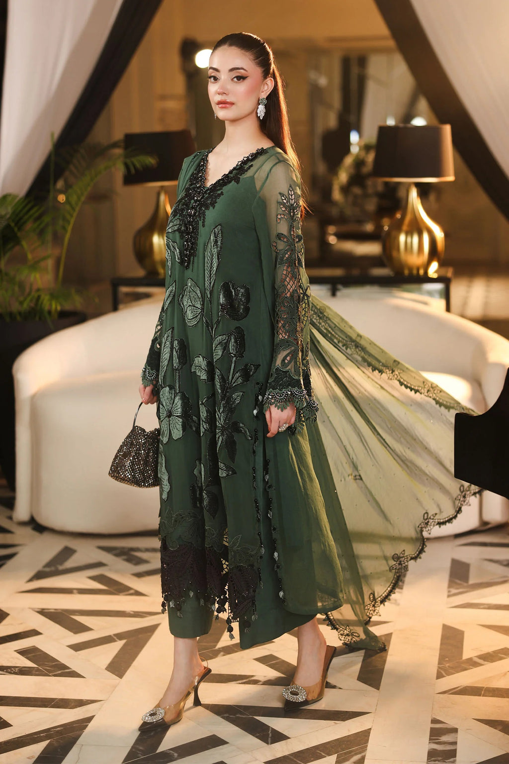 A woman models a dark green embroidered tunic dress with sheer sleeves and a matching sheer dupatta in an elegant indoor setting with a white curved sofa and patterned floor.