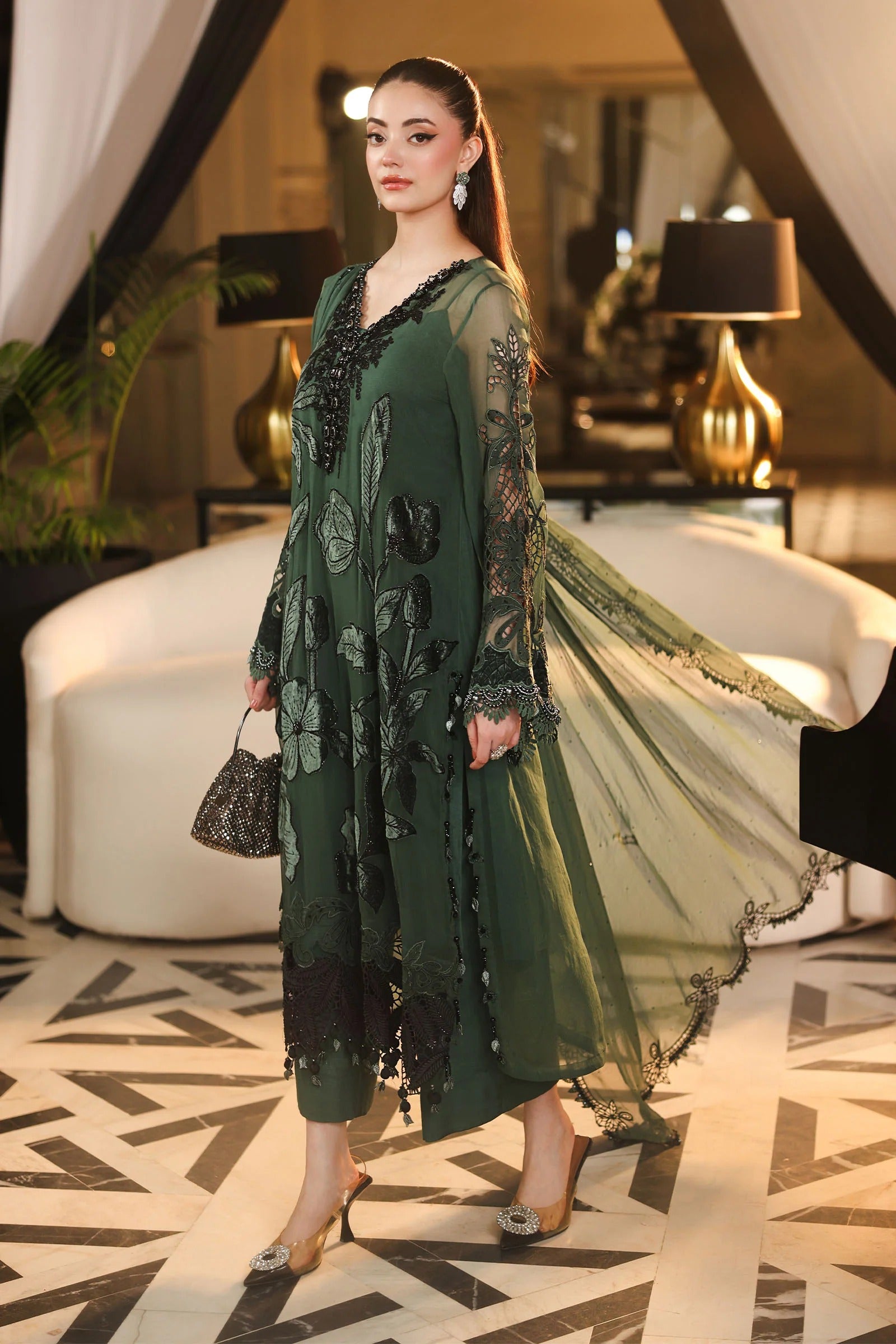A woman models a dark green embroidered tunic dress with sheer sleeves and a matching sheer dupatta in an elegant indoor setting with a white curved sofa and patterned floor.