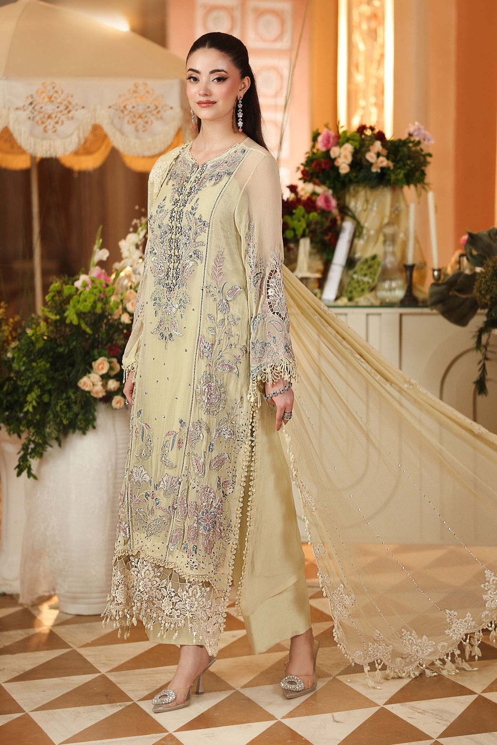 A woman models a traditional beige three-piece salwar kameez suit with intricate silver and purple embroidery, a matching sheer dupatta, and silver heels in an elegant indoor setting.