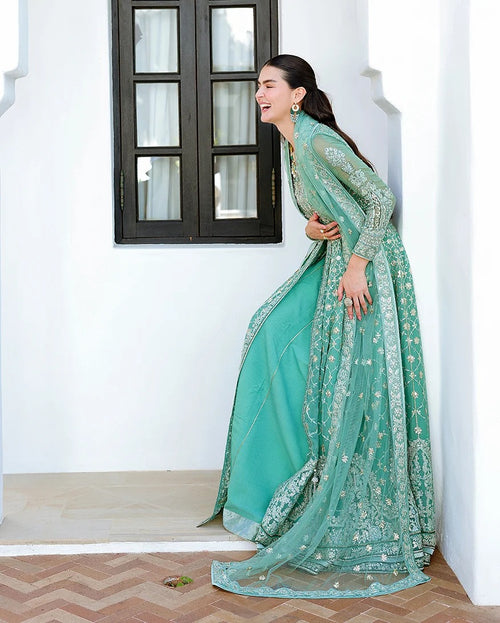 A woman wearing a mint green traditional embroidered dress with a matching dupatta, posing in a bright white-walled corridor with a dark-framed window.