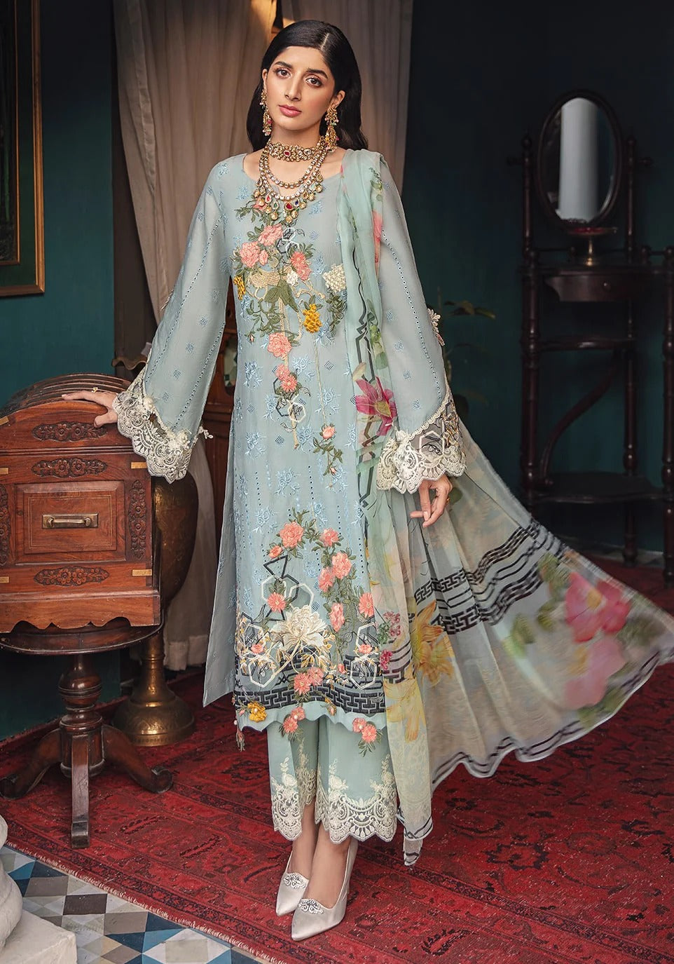 A woman models a light blue-green traditional South Asian salwar kameez suit with floral embroidery, lace-trimmed sleeves and hem, and a matching printed dupatta in an ornate room.