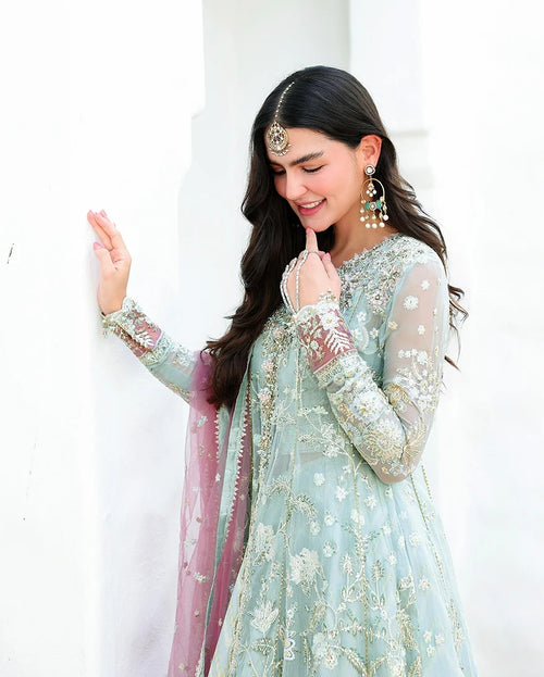 A woman wearing a traditional light blue-green embroidered kurta with a pink dupatta, gold jewelry, and a headpiece, posing against a white wall.