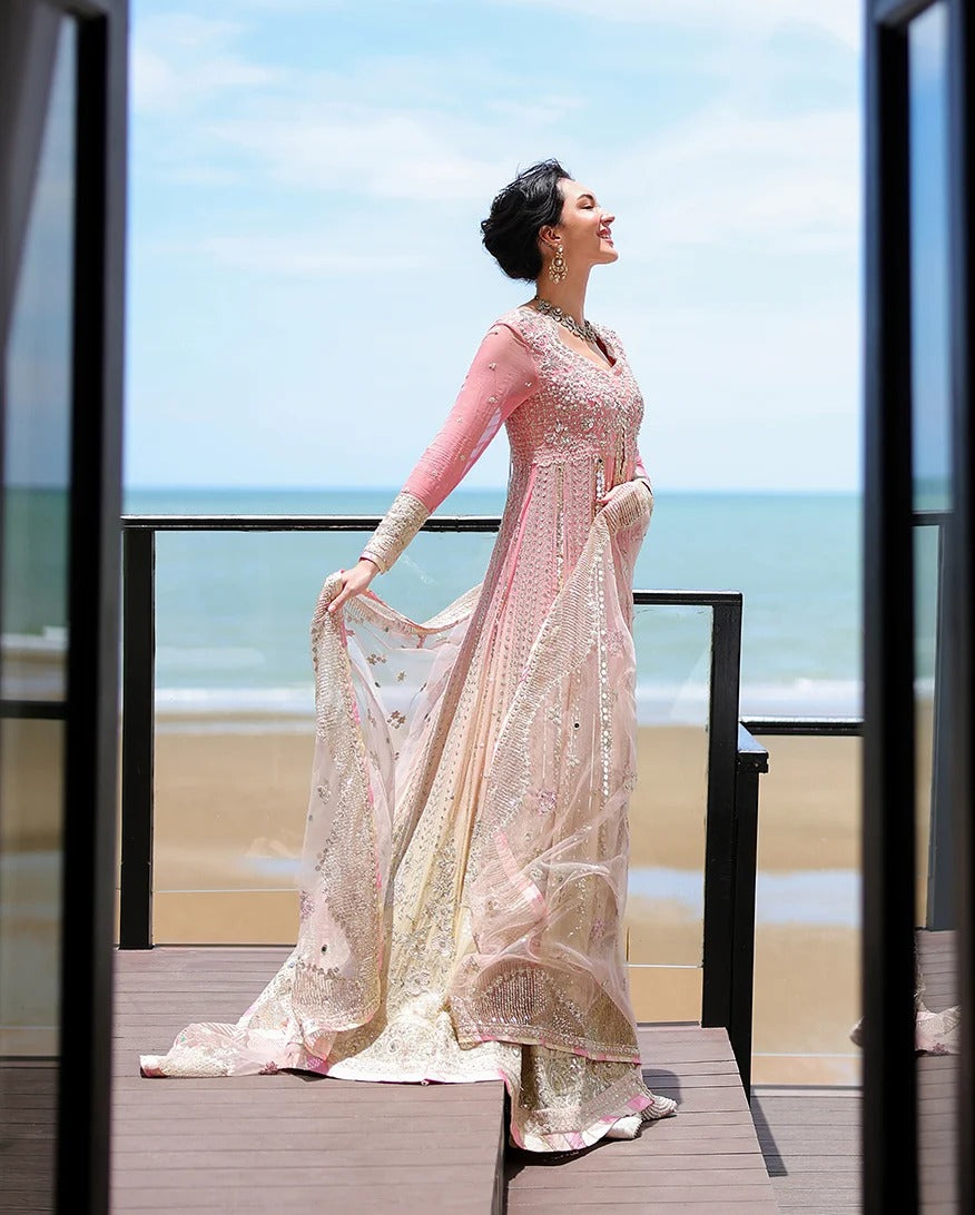 A woman wearing a pink and beige heavily embellished traditional gown stands on a wooden deck overlooking the ocean, holding out one side of the flowing dress.