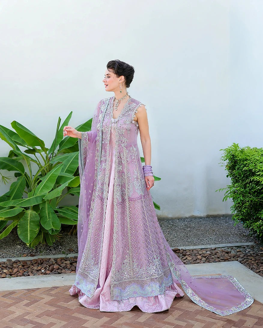 A woman models a traditional lavender pink embroidered Anarkali gown with a matching sheer dupatta, standing outdoors against a white wall with green plants.