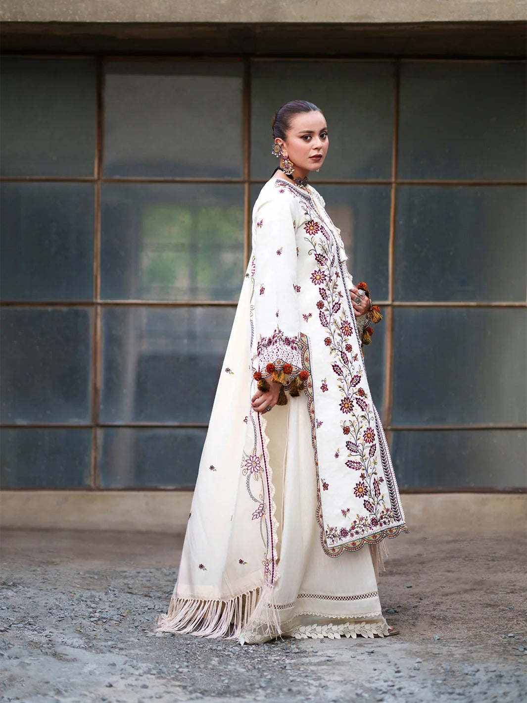 A woman models a traditional white embroidered outfit with a long coat and matching skirt, standing in front of a large windowed wall.