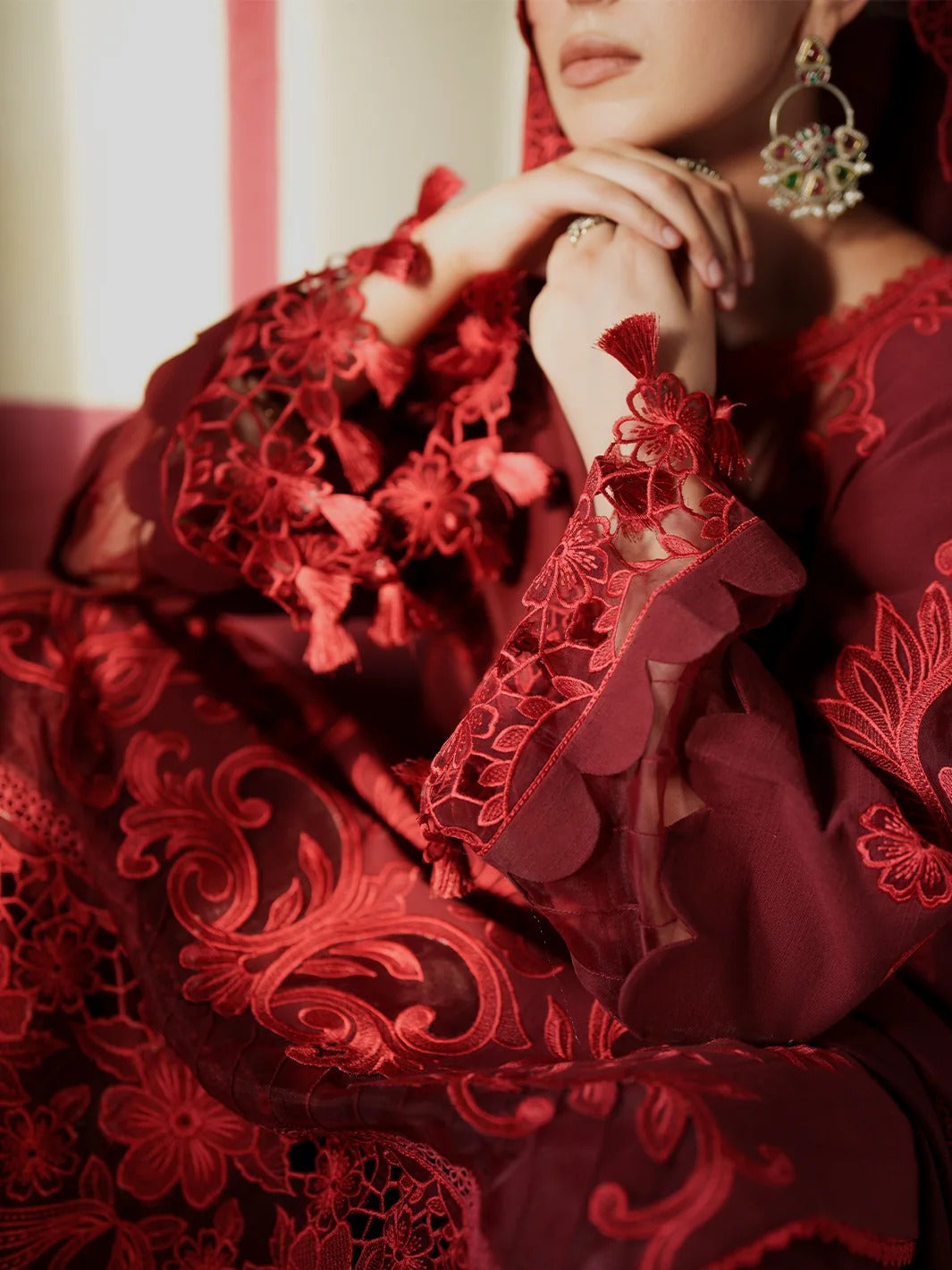 A close-up of a model wearing a rich red traditional embroidered dress with intricate lace and tassel details, paired with ornate green and gold statement earrings.