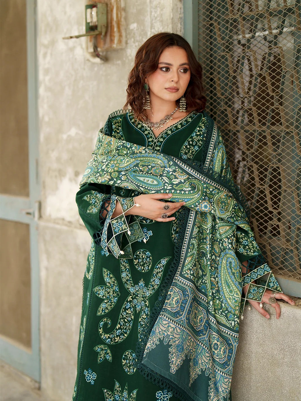A woman models a dark green traditional outfit with intricate paisley and floral patterns in light green, teal, and white, featuring a matching dupatta draped over her shoulders.