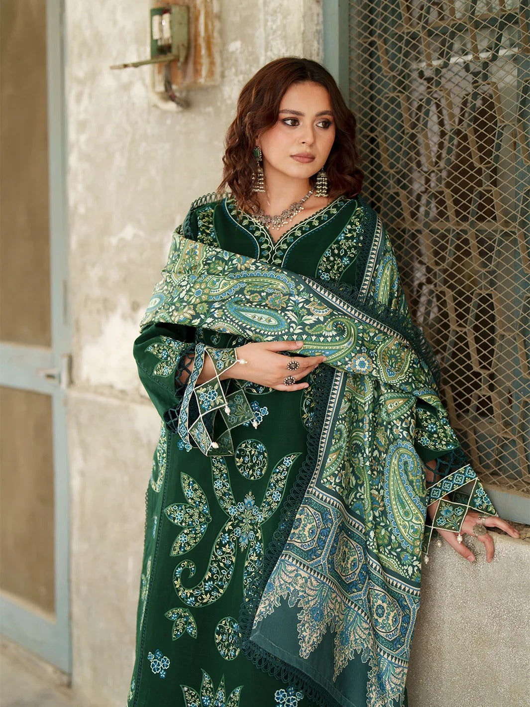 A woman models a dark green traditional outfit with intricate paisley and floral patterns in light green, teal, and white, featuring a matching dupatta draped over her shoulders.