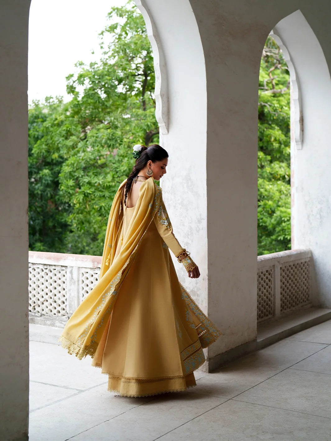 A woman wearing a flowing mustard yellow traditional outfit with a matching dupatta walks through a covered walkway framed by white arches and green trees in the background.