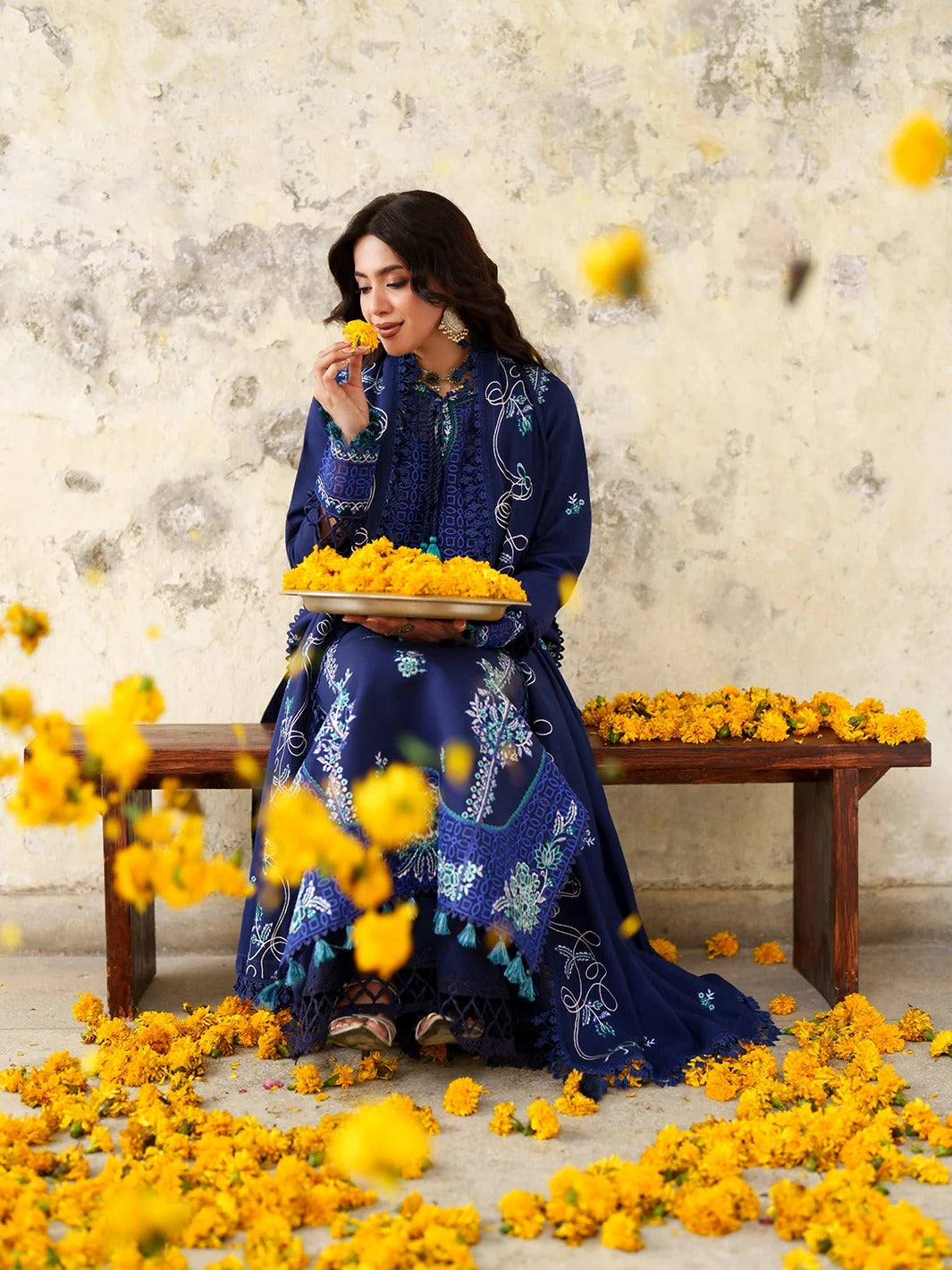 A woman in a deep blue embroidered traditional dress sits on a wooden bench, holding a tray of yellow marigold flowers against a textured beige wall with scattered yellow flowers on the floor.
