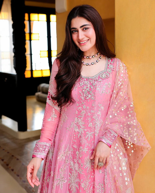 A woman wearing a pink traditional embroidered dress with a matching sheer dupatta, decorated with silver floral embroidery and mirror work, standing in an indoor setting with a warm yellow wall background.