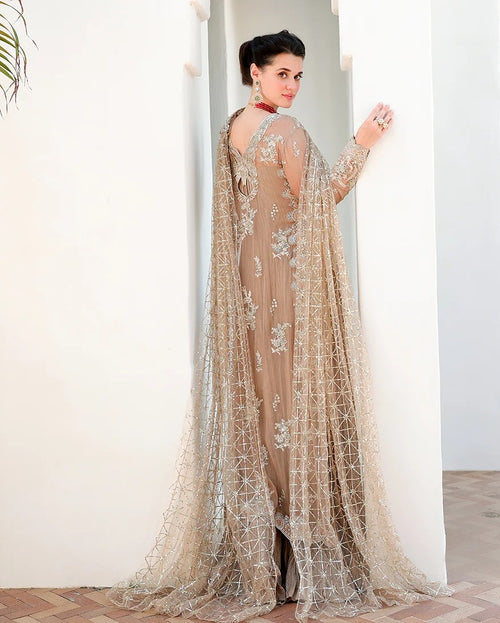 A woman wearing a beige embellished traditional gown with a sheer embroidered cape, standing in a doorway and looking back over her shoulder.