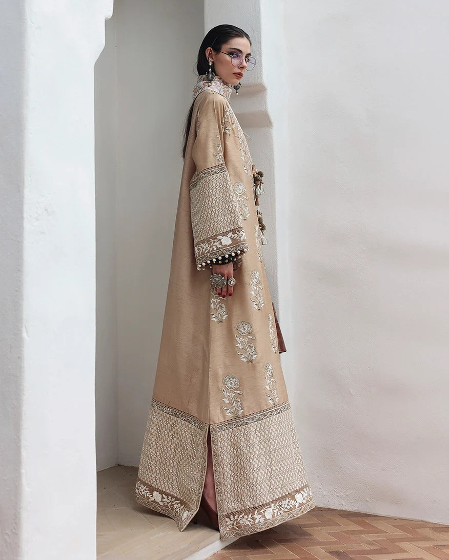 A woman modeling a long beige embroidered traditional dress with wide sleeves and ornate metallic detailing, standing in a white architectural setting with a wood floor visible at the bottom.