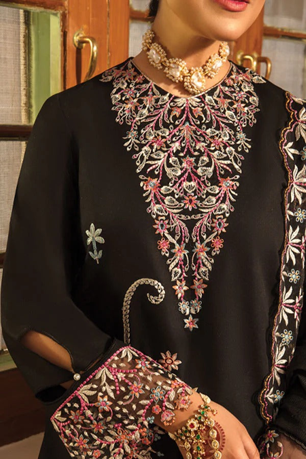 A close-up of a woman wearing a black traditional embroidered outfit with a large floral neckline motif in pink, white, and metallic thread. She is also wearing a chunky gold-toned necklace and holding a matching embroidered clutch or pouch.
