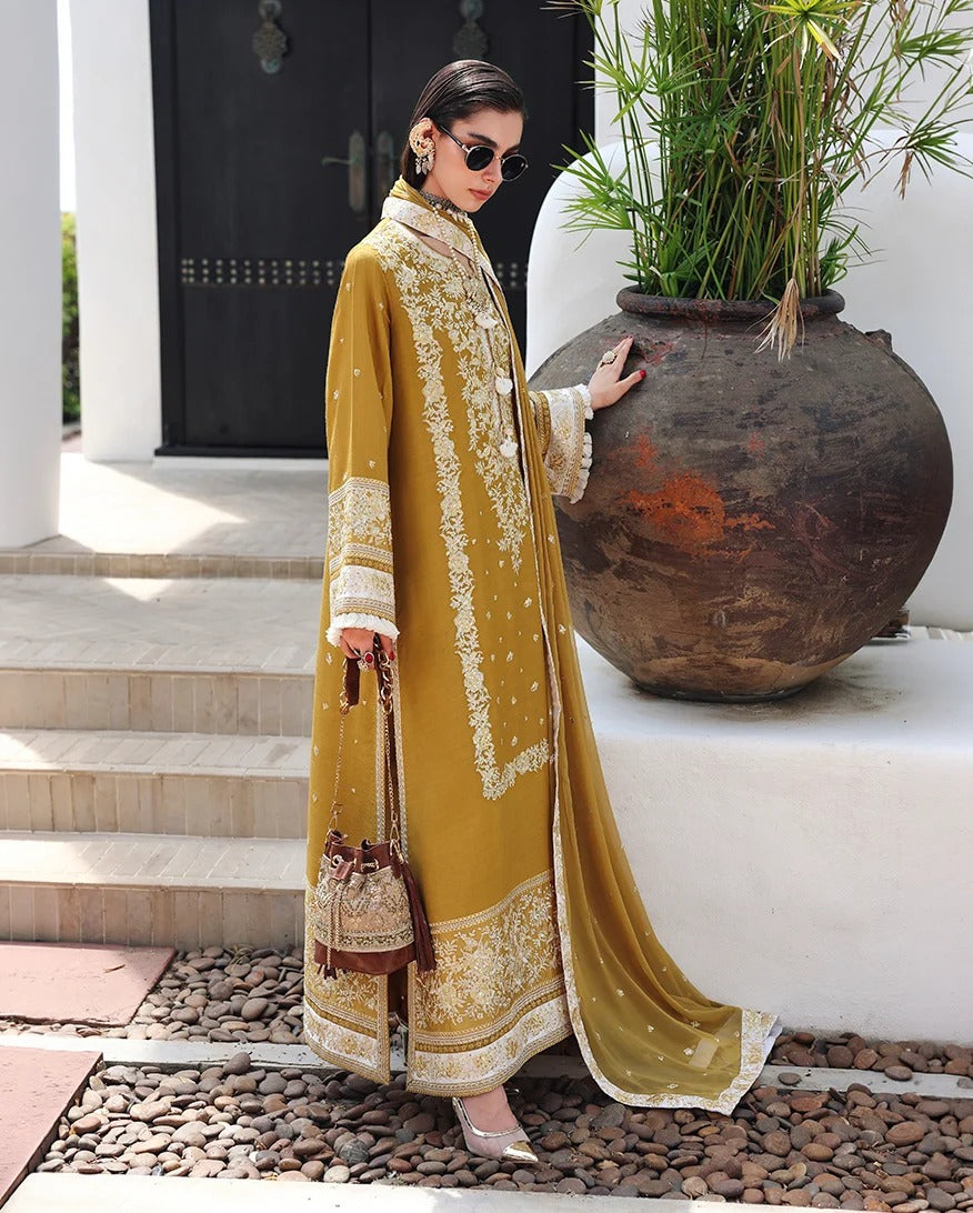 A woman models a long, flowing mustard yellow embroidered outer garment with white and gold detailing, standing outdoors next to a large brown planter pot.