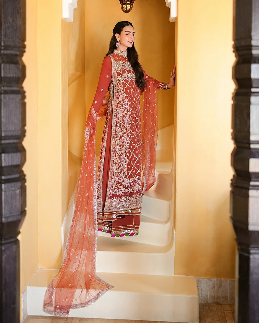 A woman models an ornate peach and red embroidered traditional outfit with a matching sheer dupatta, standing on white stairs against a warm yellow wall.