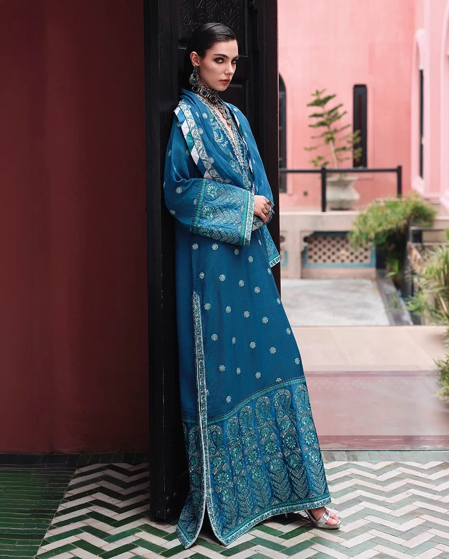 A woman models a traditional blue embroidered kurta set in an outdoor courtyard setting with a pink wall and patterned tile floor.