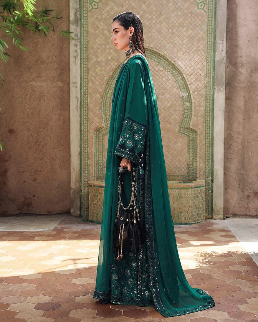 A woman stands in profile wearing an elegant emerald green traditional outfit with intricate gold embroidery and a matching sheer draped overlay, set against a Moroccan-style tiled courtyard wall.