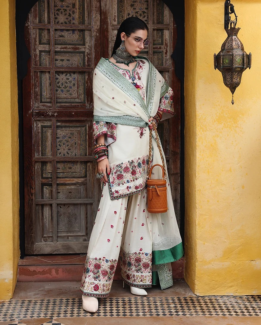 A woman models a traditional South Asian outfit consisting of an off-white salwar suit with colorful floral embroidery, a matching dupatta, and a green inner lining, standing in front of a rustic wooden door and a yellow wall.