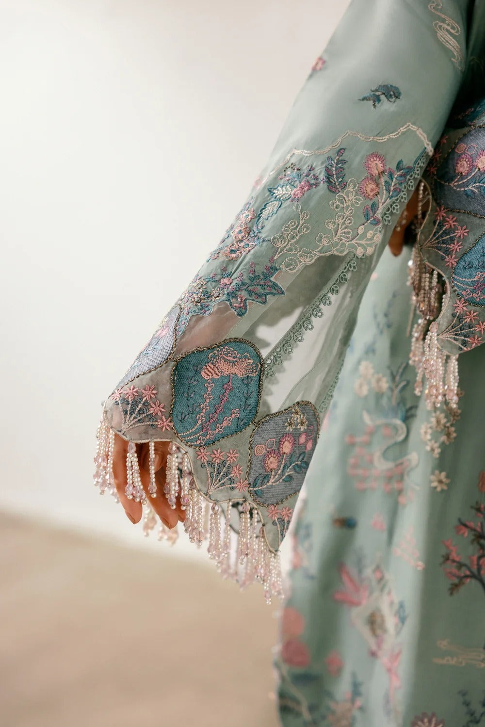 A close-up of a person wearing a flowing mint green embroidered garment with ornate floral and bird motifs, beaded fringe along the sleeve edge, and a visible hand holding the fabric.