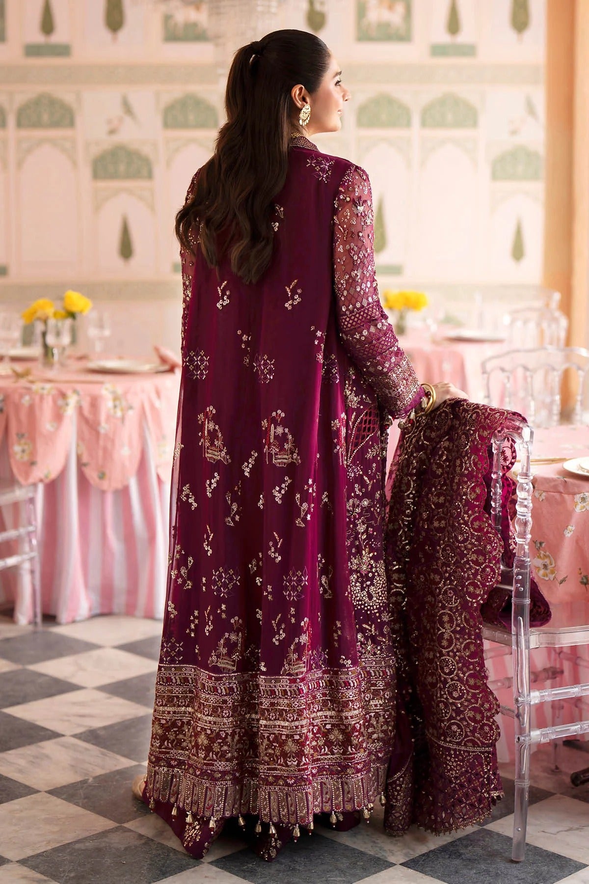 A woman stands in a decorated indoor event space wearing a long magenta-purple embroidered traditional outfit with a matching draped dupatta. The outfit features intricate gold and silver mirror work and floral embroidery, particularly dense around the border and sleeves.