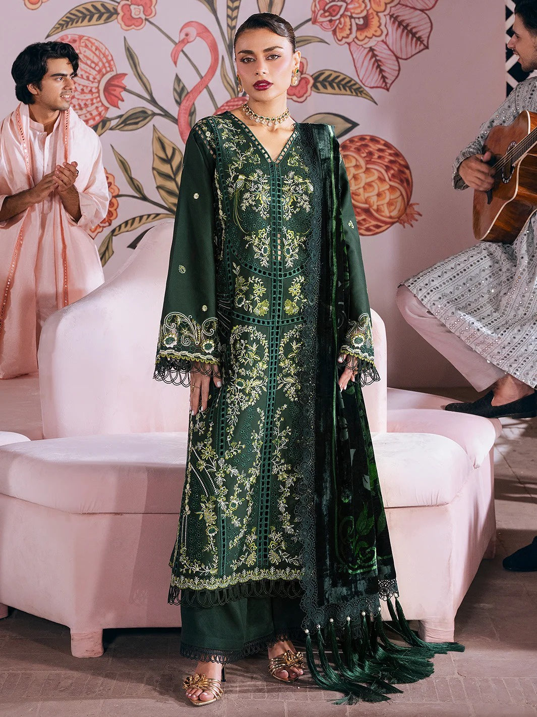 A woman models a dark green traditional embroidered outfit with gold detailing, paired with a matching dupatta, in a styled indoor setting with pink furniture and other people partially visible in the background.