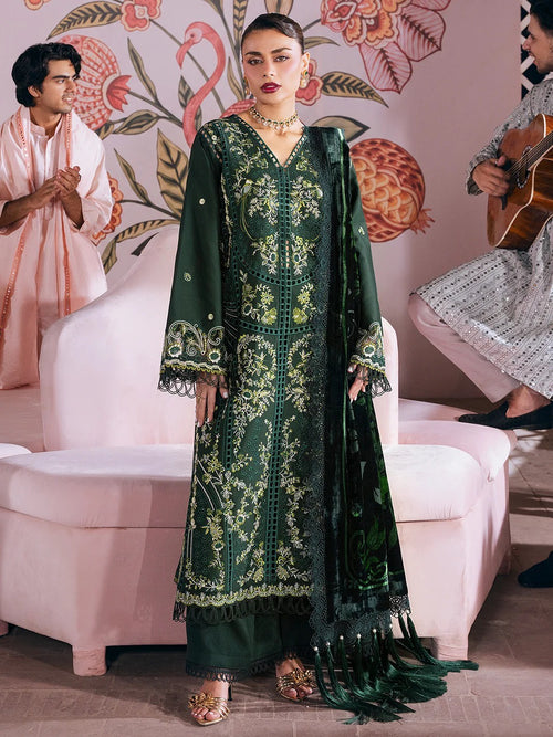 A woman models a dark green traditional embroidered outfit with gold detailing, paired with a matching dupatta, in a styled indoor setting with pink furniture and other people partially visible in the background.