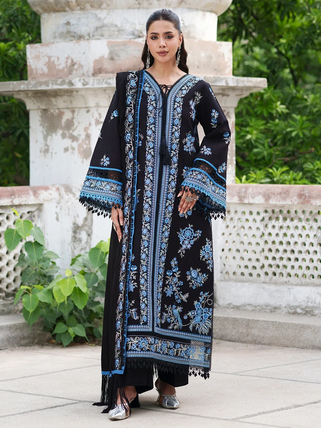 A woman models a long black embroidered kurta with intricate blue and light blue floral patterns, standing outdoors in front of a stone structure and green foliage.