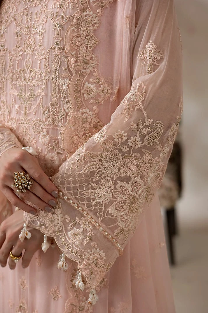 A close-up of a person wearing a light blush pink traditional embroidered outfit with sheer lace sleeves, floral embroidery, and pearl-like bead tassels. The person is also wearing ornate gold rings.