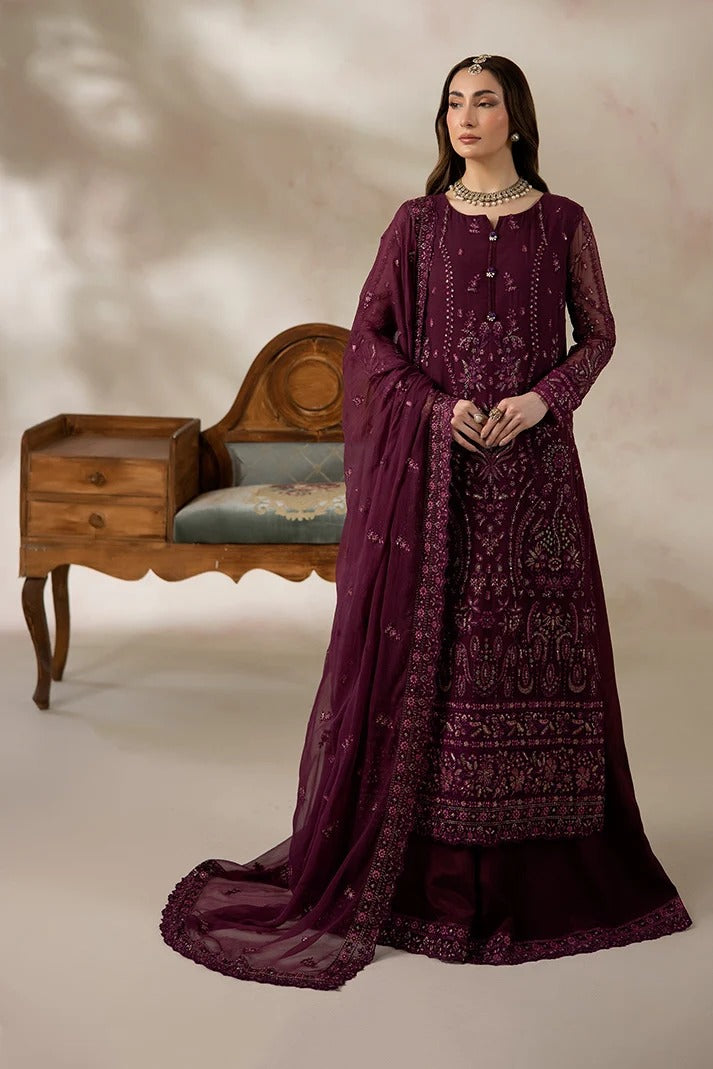 A woman models a deep purple embroidered traditional outfit with a matching sheer dupatta, standing in front of a beige studio backdrop next to a wooden bench.