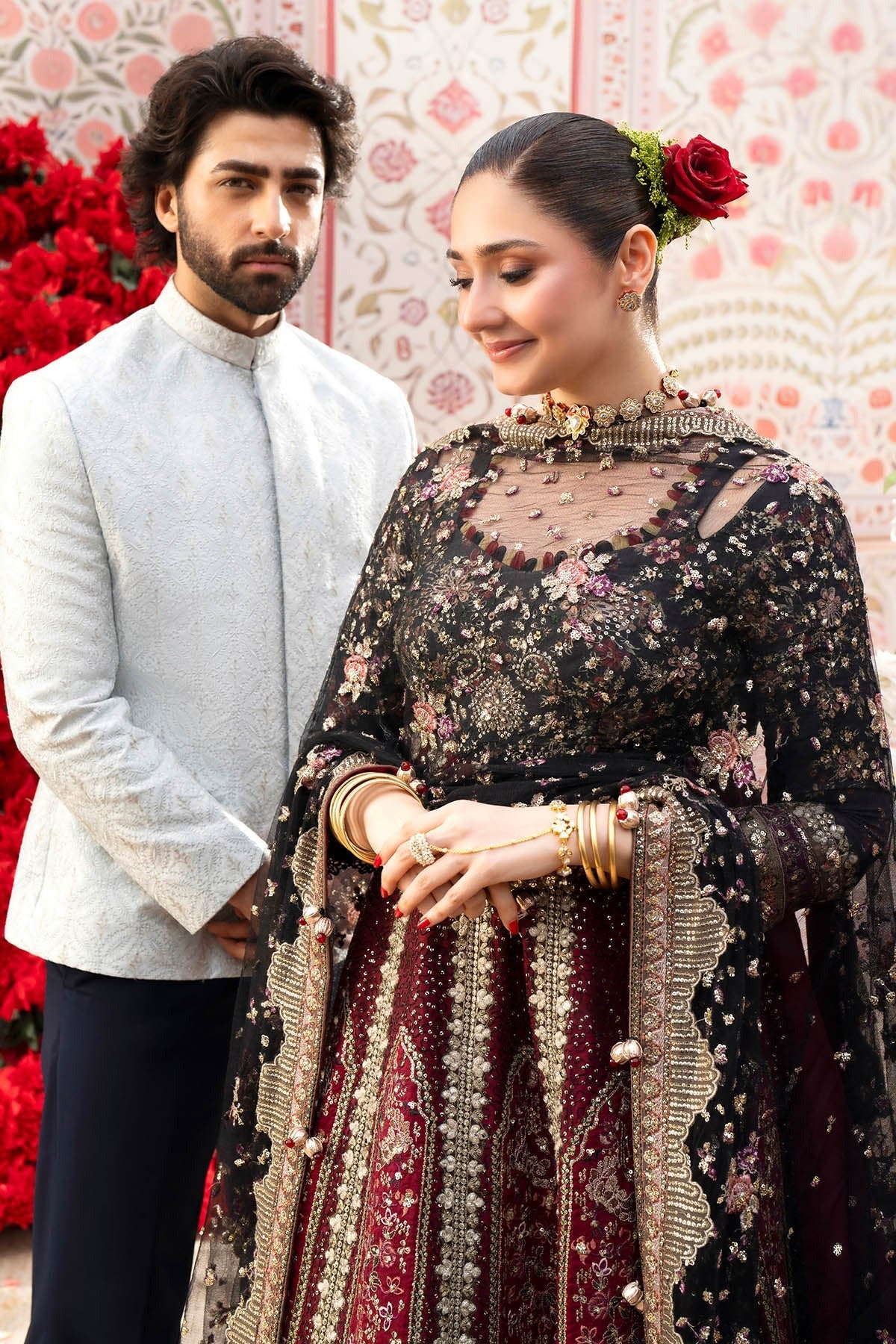 A man and a woman posing in traditional South Asian wedding attire. The woman wears an intricate black and deep maroon embroidered lehenga with gold embroidery, a sheer netted bodice, and a red rose in her hair.