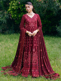 A woman stands outdoors on grass wearing a long burgundy embroidered Anarkali-style gown with a matching sheer draped overlay and tassel details. The background shows green foliage and lawn.